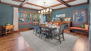 A dining area with a wooden table, chairs, and a chandelier. There is a window with curtains and a bench with cushions. - Walk to Beach & Pier | Fully-Loaded Kitchen (Sheboygan)