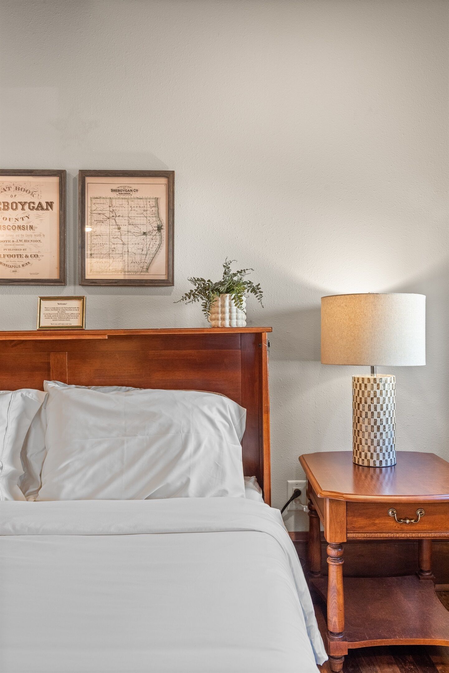 A neatly made bed with white linens, a wooden nightstand with a lamp, and two framed maps on the wall.