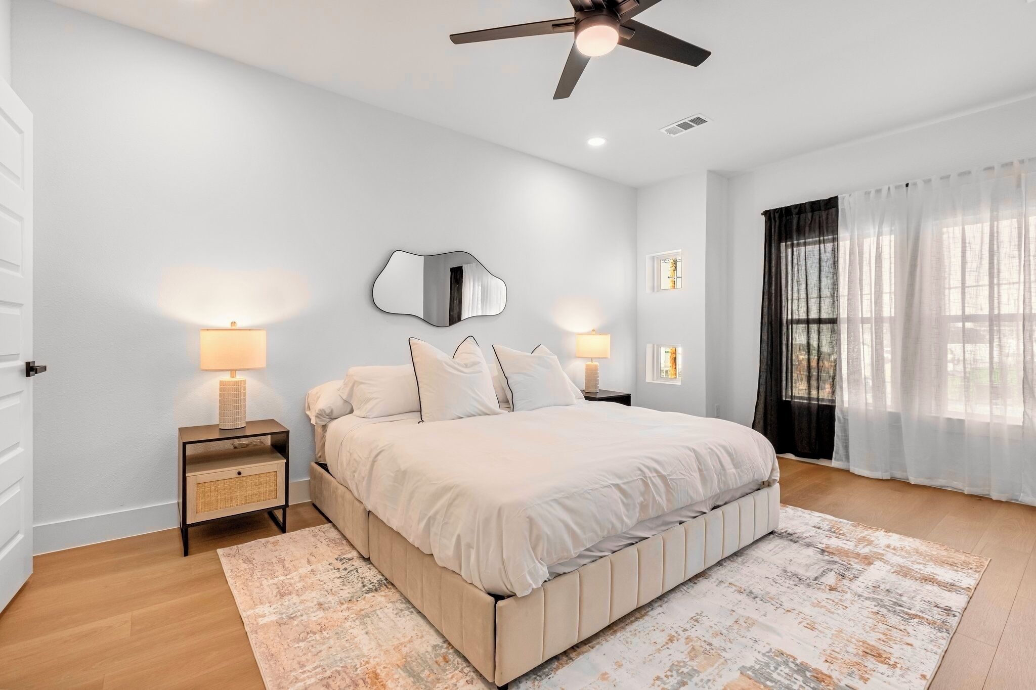 A bedroom with a bed, two bedside tables with lamps, a ceiling fan, a large mirror, and a window with sheer curtains.