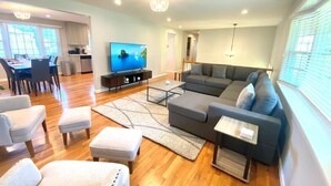 Ein modernes Wohnzimmer mit einer großen Eckcouch, einem Essbereich mit Tisch und Stühlen, einem Fernseher auf einem Lowboard und einem Kronleuchter.