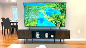 Ein modernes Wohnzimmer mit einem Flachbildfernseher auf einem dunklen Holz-TV-Schrank, einem Essbereich mit Tisch und Stühlen sowie einem grauen Fliesenboden.