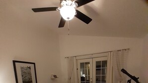 Quarto com cama, ventilador de teto, telescópio e porta com painéis de vidro.