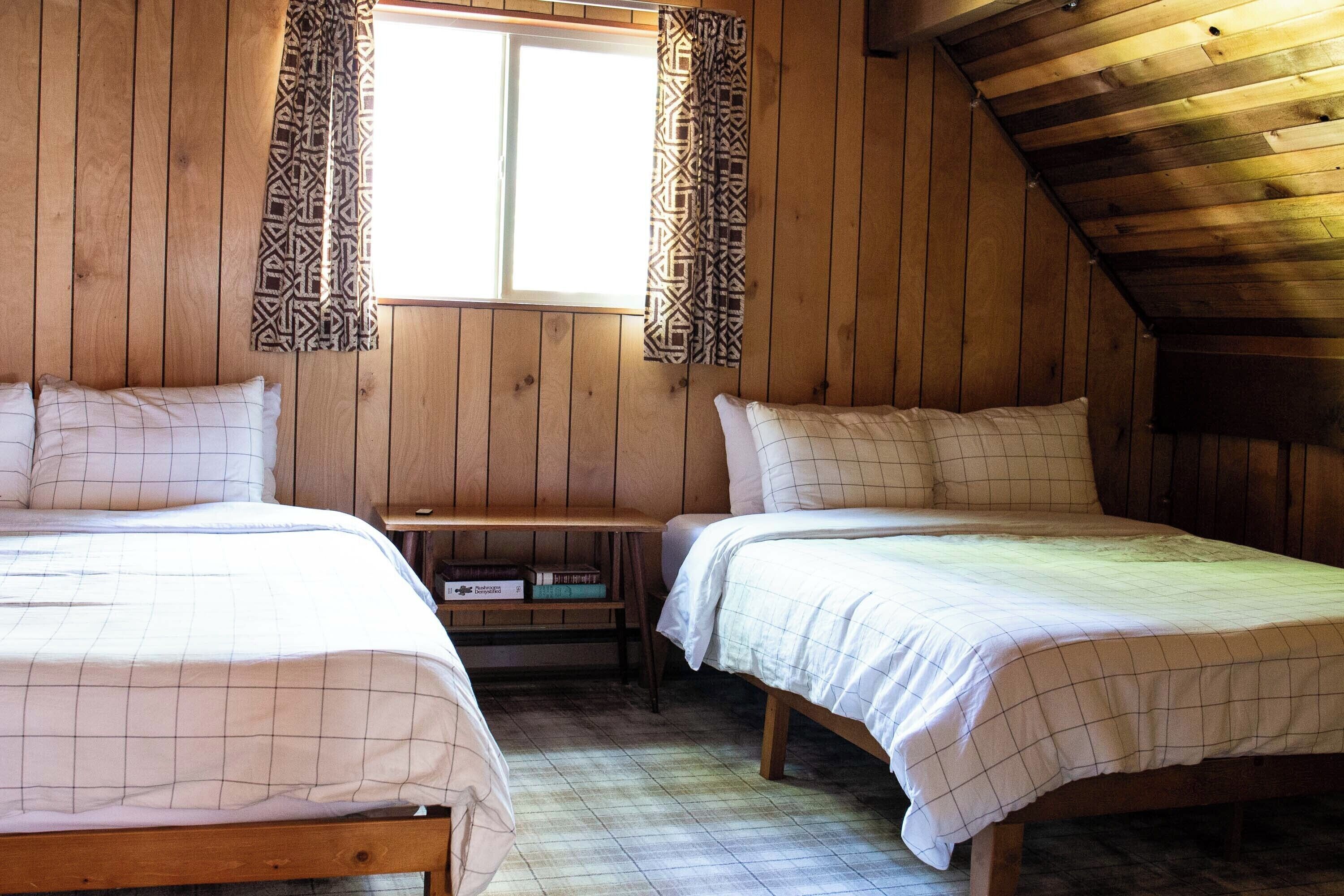 A room with two beds, wooden walls, a window with patterned curtains, and a sloped ceiling.