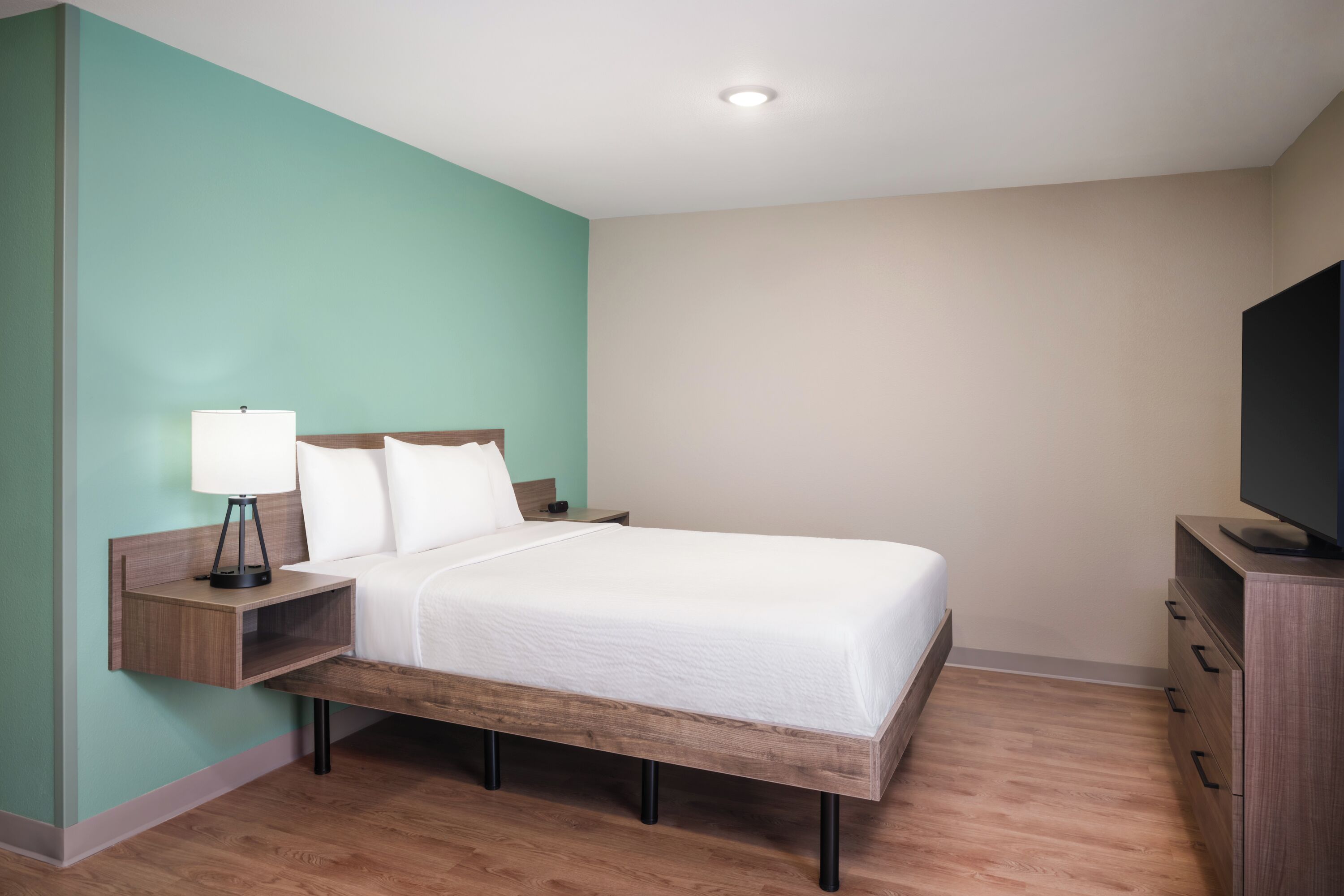 Habitación de hotel ordenada con cama, mesita de noche, lámpara y televisor sobre un aparador.