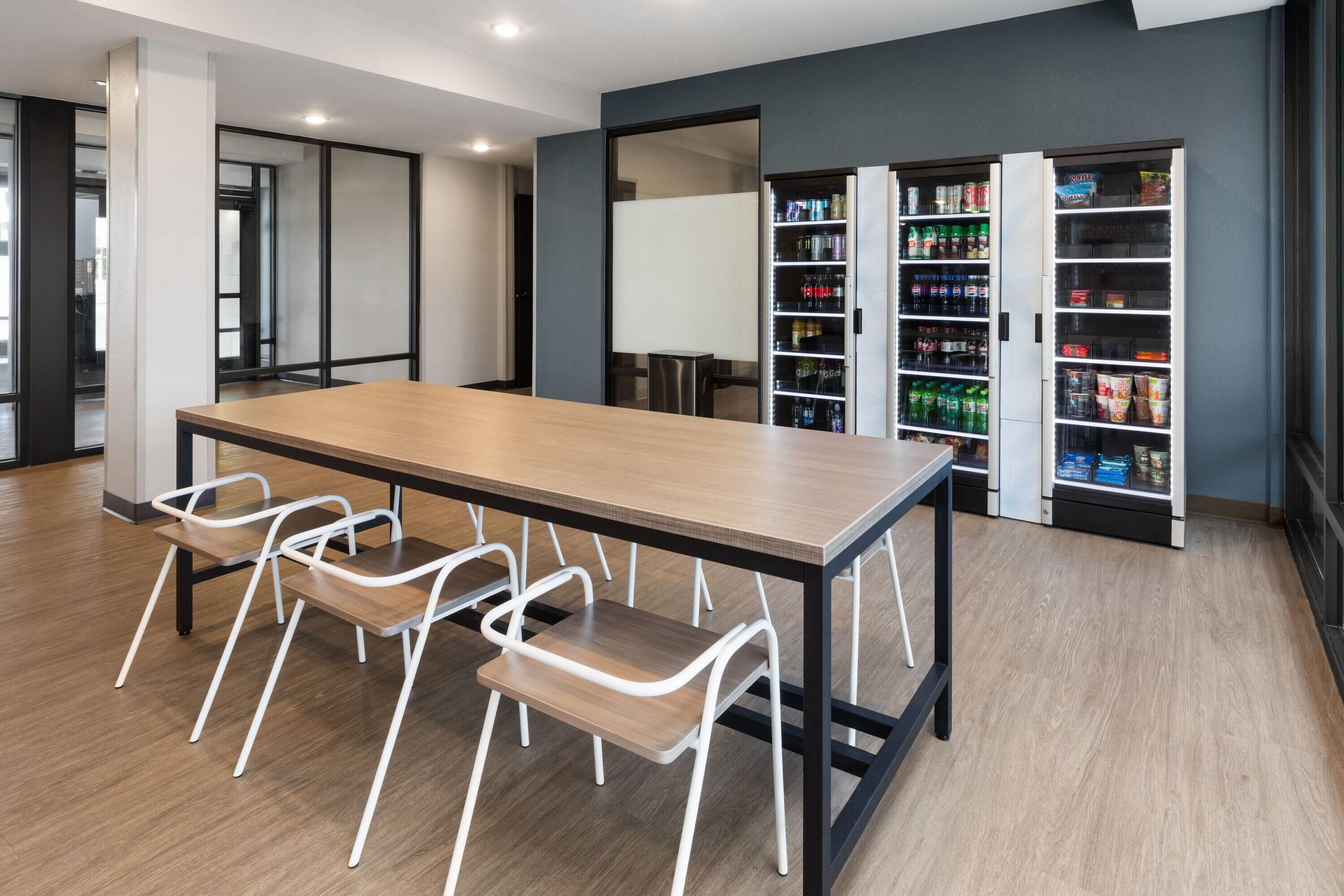 A modern meeting room with a long table and four white chairs, a whiteboard, and a vending machine stocked with beverages.