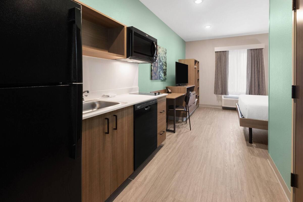 A compact hotel room with a kitchenette, a bed, a TV, and a window with curtains.