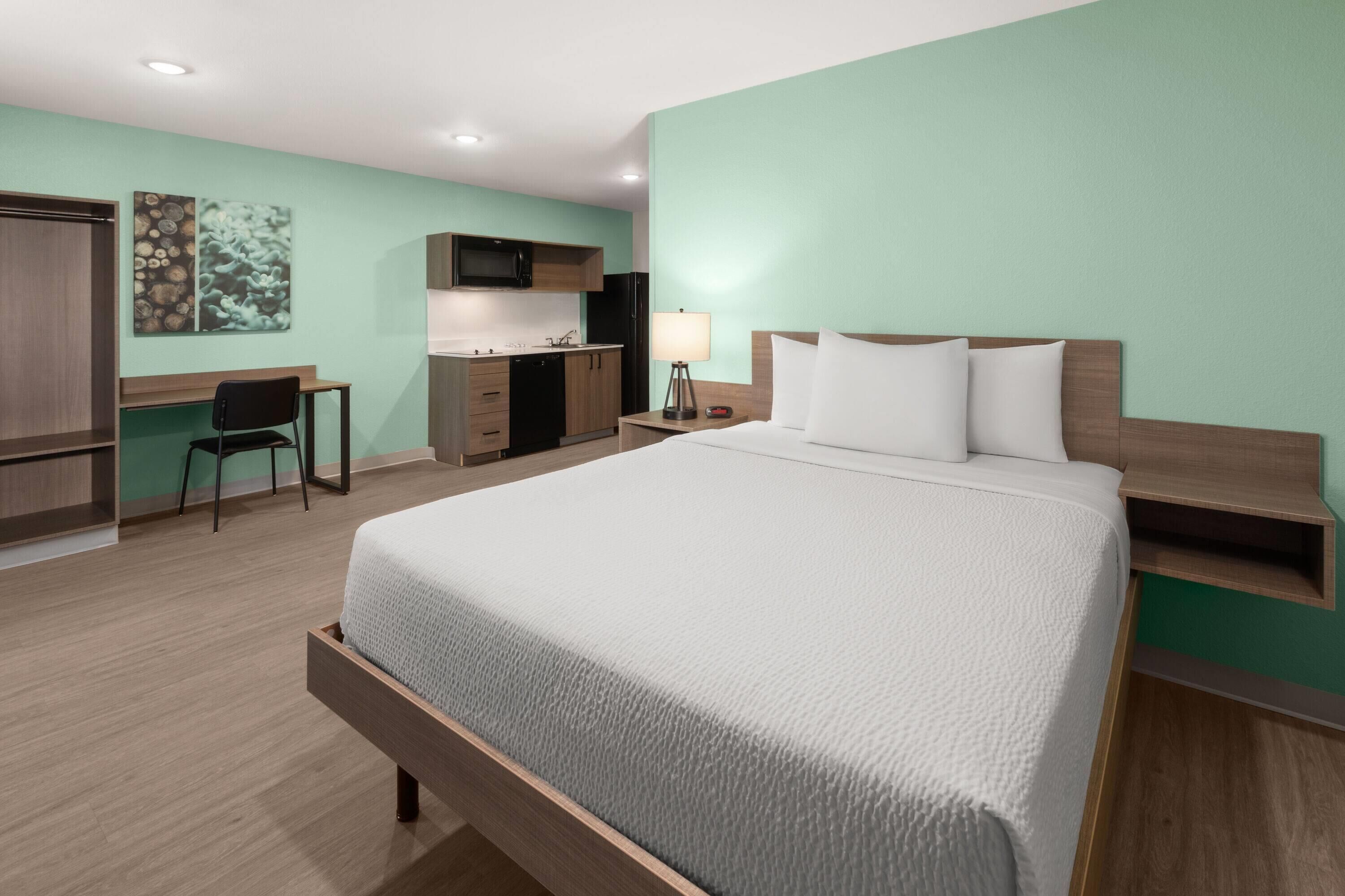 A hotel room with a bed, desk, chair, kitchenette, and a nightstand with a lamp.