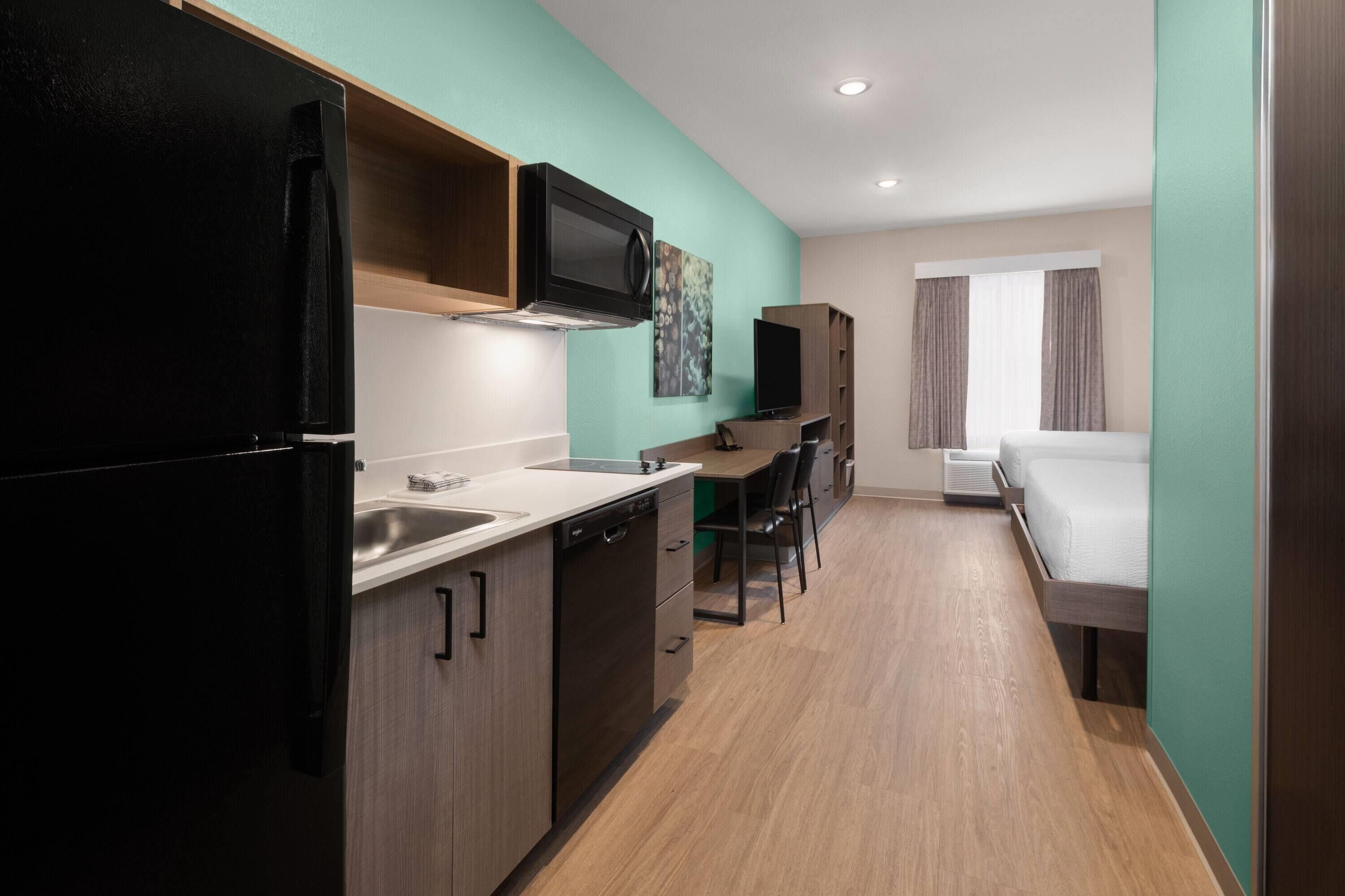 A compact hotel room with a kitchenette, a bed, a desk with a chair, and a television.