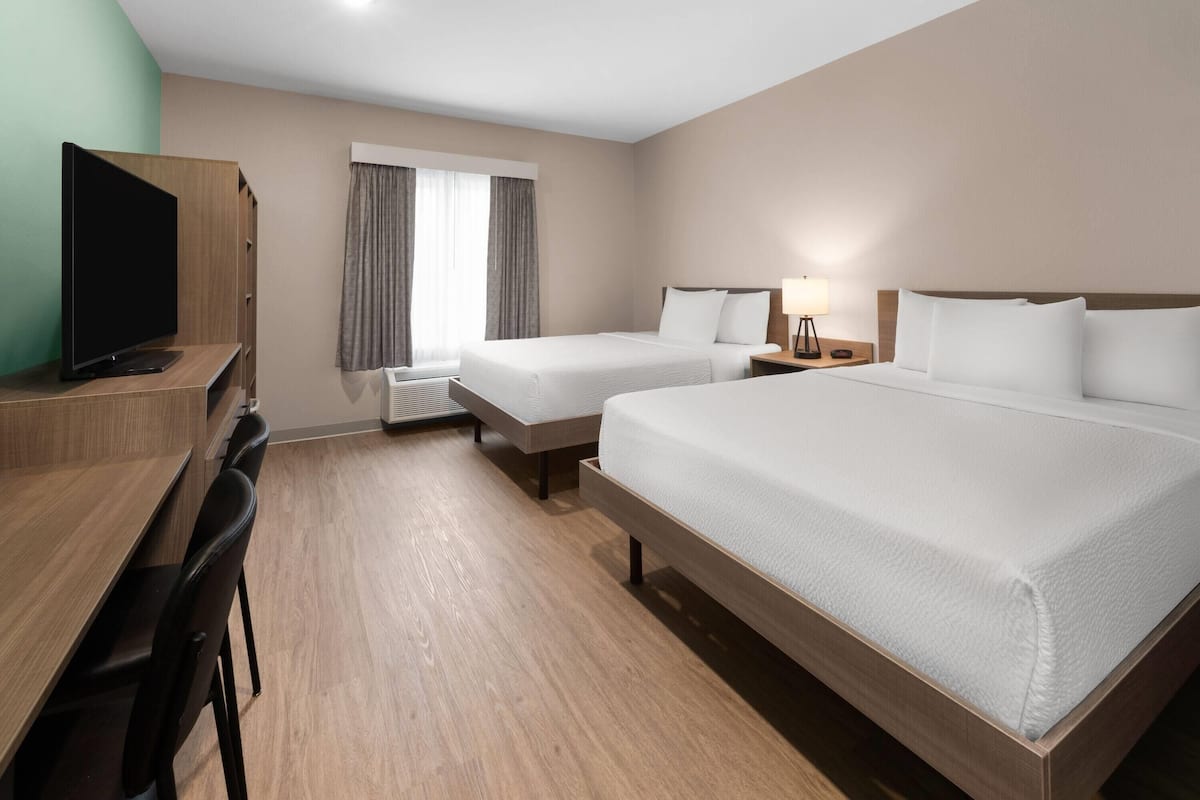 A hotel room with two beds, a desk with chairs, a television, and a window with curtains.