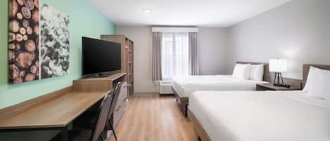 Une chambre d’hôtel avec un bureau en bois, une télévision, deux lits, une fenêtre avec des rideaux et des œuvres d’art accrochées au mur.