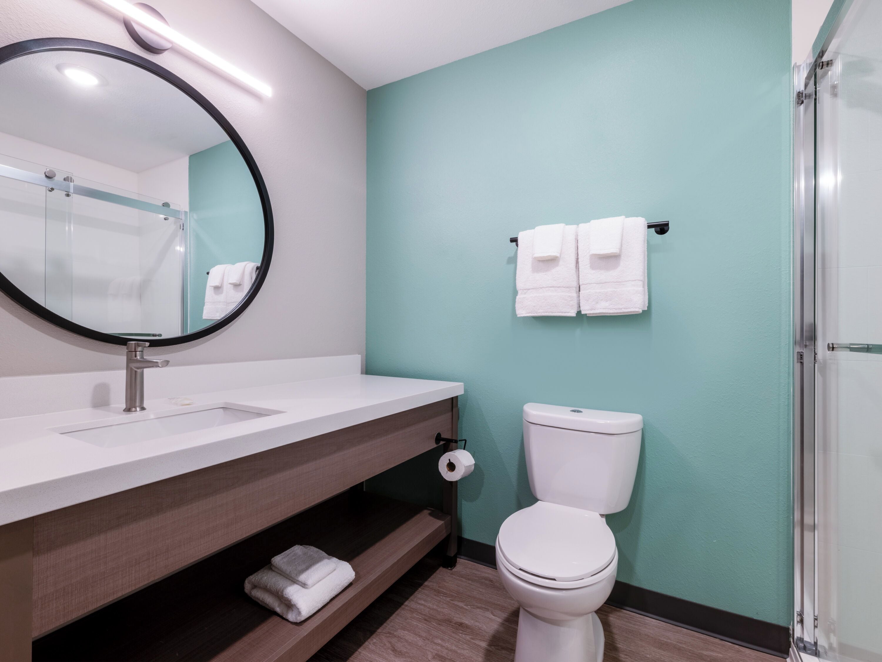 A modern bathroom with a white sink, a round mirror, a toilet, and a glass shower enclosure.