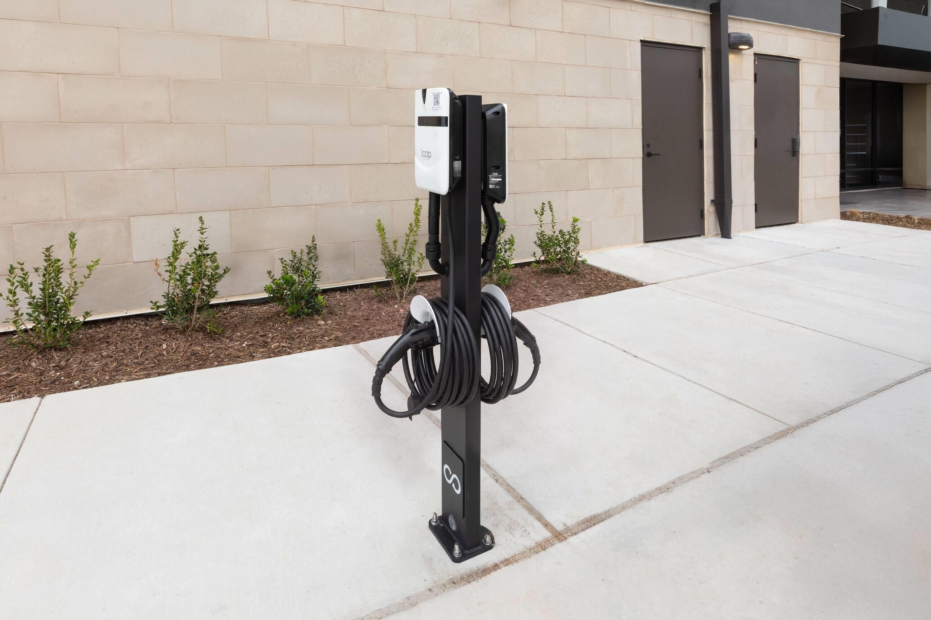 Une borne de recharge pour véhicules électriques, avec un câble enroulé autour du poteau, située devant un bâtiment dont le mur est en briques et qui possède une porte.