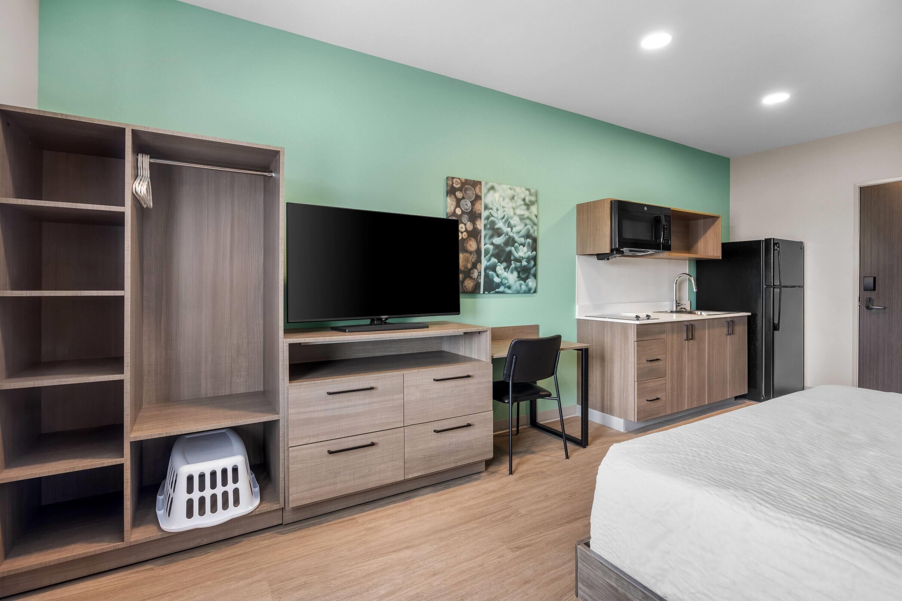 Una habitación de hotel moderna con cama, televisión, escritorio con silla, armario y kitchenette.