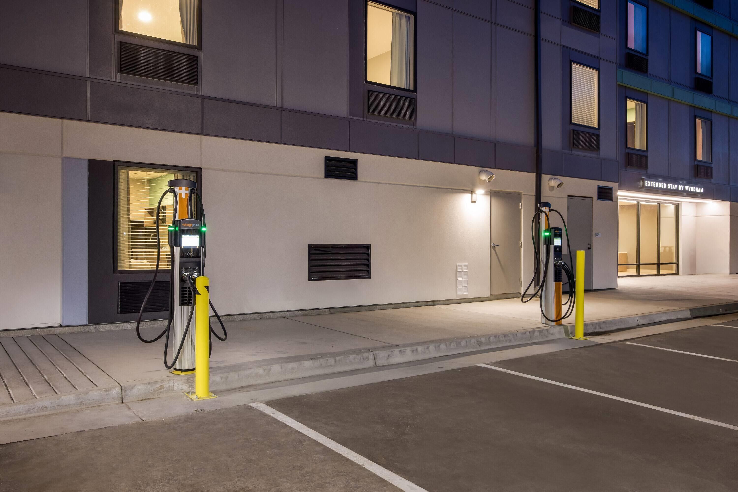 Un estacionamiento nocturno con estaciones de carga para vehículos eléctricos, entrada a un edificio y un cartel que indica 'Extended Stay by Wyndham'.