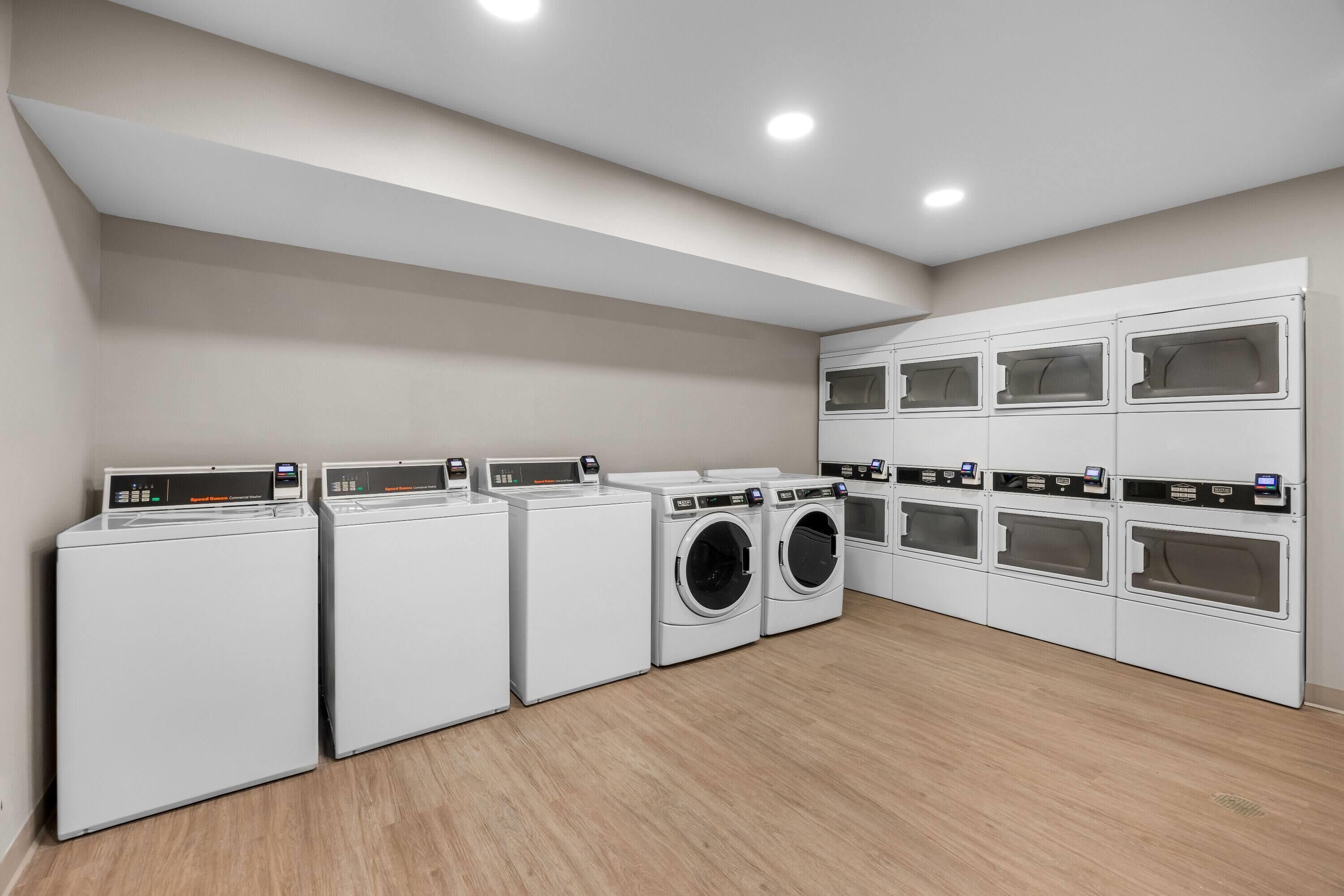 Une buanderie équipée de plusieurs machines à laver et de plusieurs sèche-linge, avec un plancher en bois et un plafond blanc doté d’un éclairage encastré.