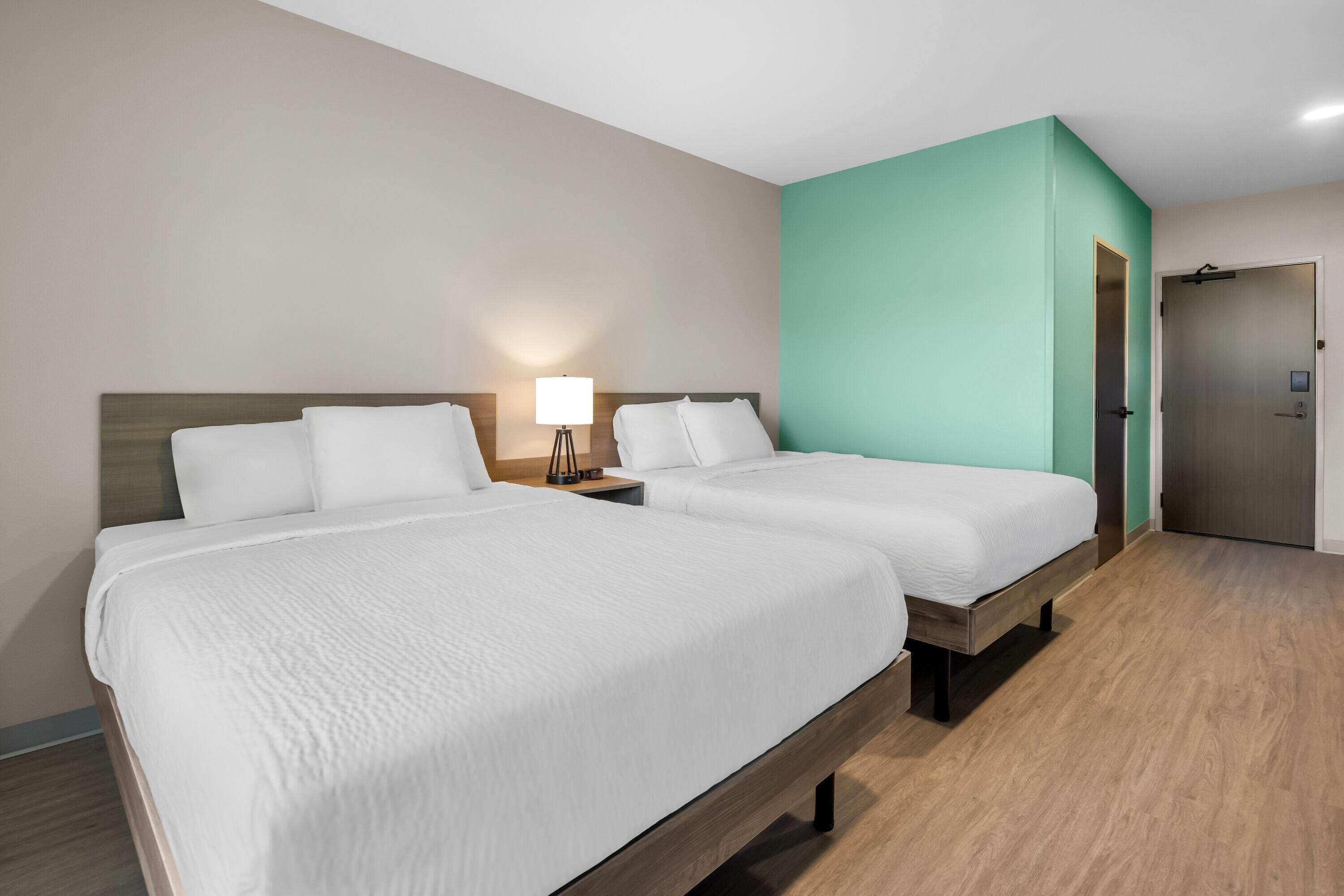 Habitación de hotel con dos camas, una mesita de noche con lámpara y una puerta al baño.