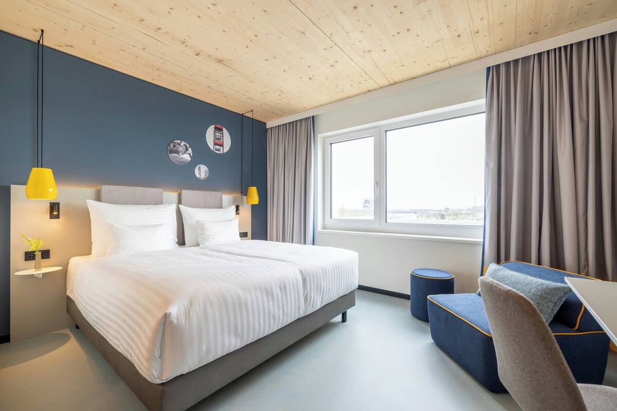 A modern hotel room with a large bed, bedside lamps, a small table, and a view of the city through the window.
