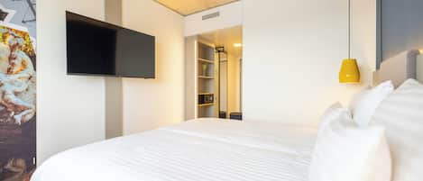 A hotel room with a large bed, a mounted TV, a yellow pendant light, and a closet with shelves.