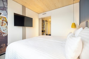 Une chambre d’hôtel avec un grand lit, un téléviseur fixé au mur, un luminaire suspendu jaune et une armoire avec des étagères.