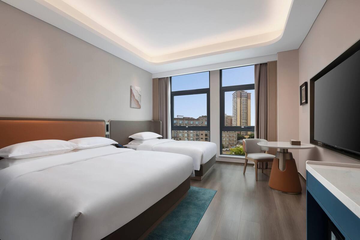 A modern hotel room with two beds, a desk, a chair, a TV, and large windows offering a city view.