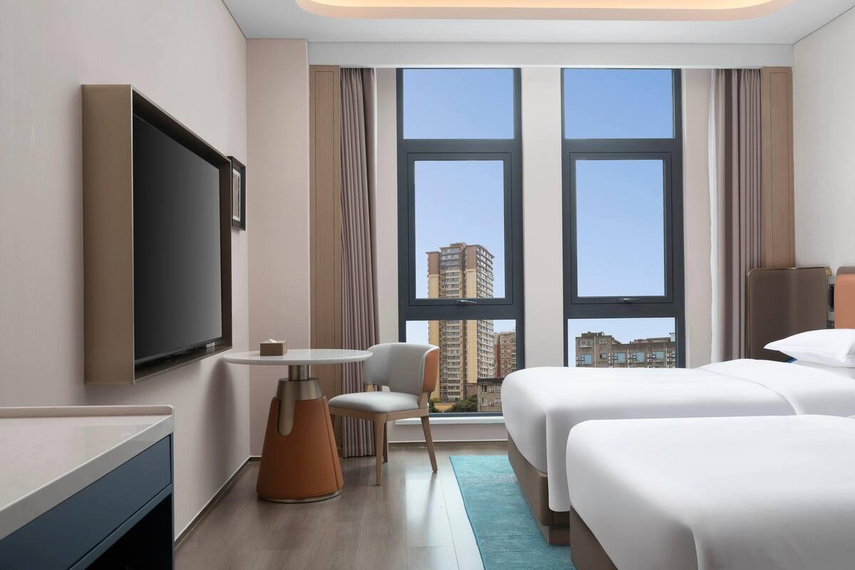 A hotel room with a large TV, a desk, a chair, two beds, and a view of tall buildings through large windows.
