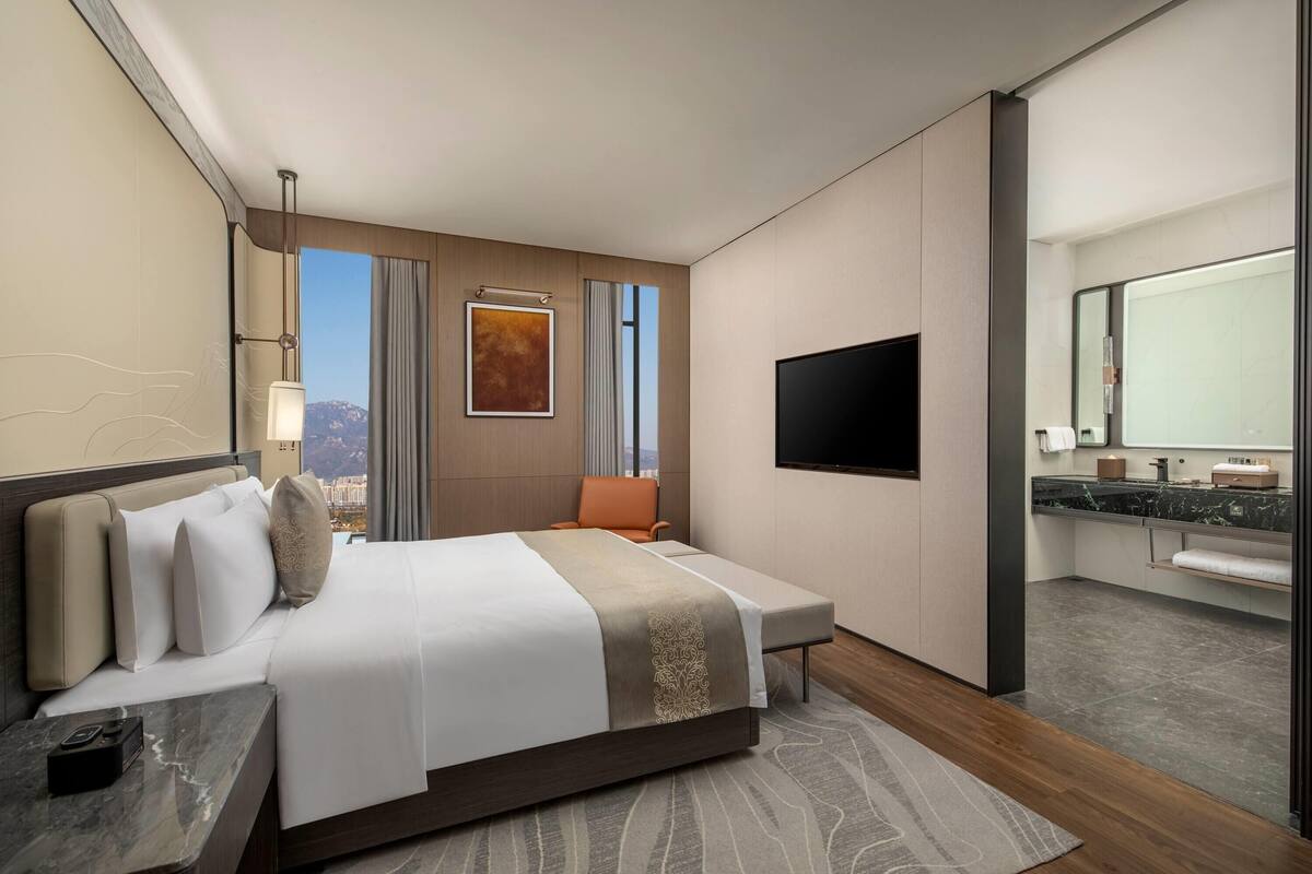 A modern hotel room with a large bed, a flat-screen TV, a bathroom with a marble countertop, and a city view.