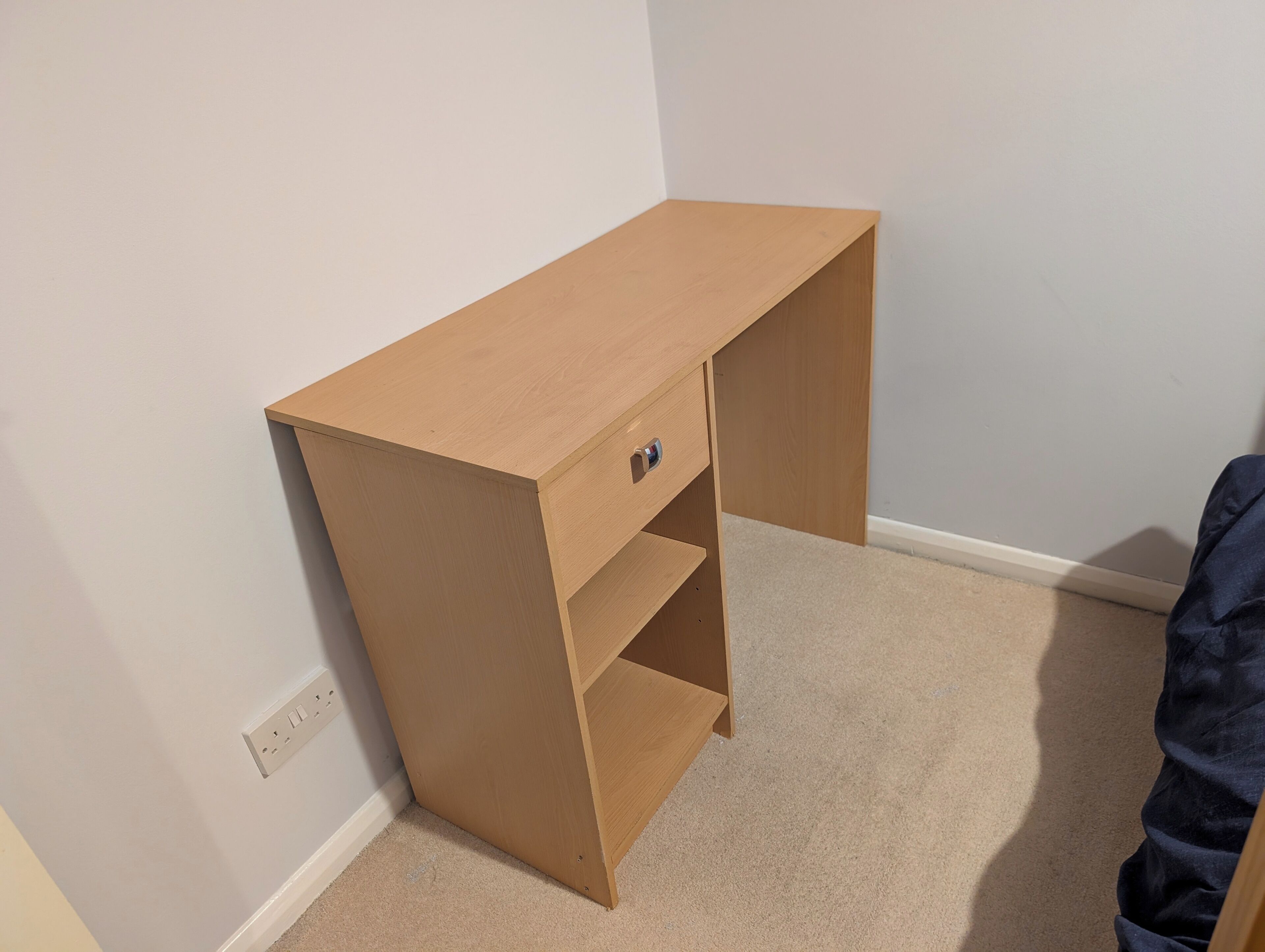 A corner bedroom with a wooden desk, a single drawer, and a carpeted floor.