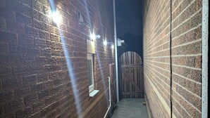A narrow brick corridor with a wooden gate at the end, a yellow hose on the left, and a light fixture on the wall. - Spacious 3 bedroom Family house with games room in quiet area (Braunstone Town)