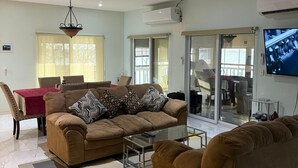 Ein modernes Wohnzimmer mit einer braunen Sofagarnitur, einem Glastisch und einem roten Tisch mit Stühlen. Ein großes Fenster mit leichten Vorhängen, eine Klimaanlage an der Wand und ein Flachbildfernseher, der über einem Glastisch montiert ist.