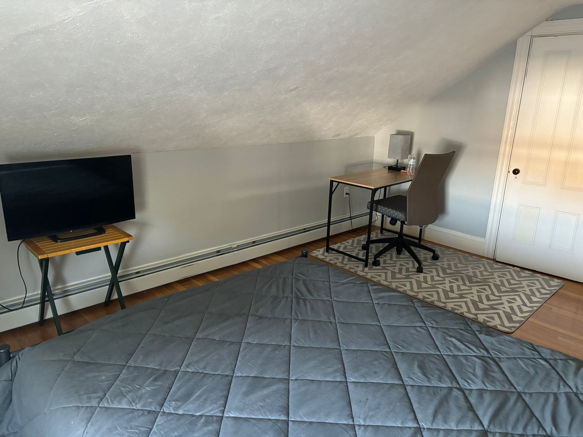 Quarto pequeno e acolhedor com televisão sobre suporte de madeira, escrivaninha com cadeira e tapete estampado no chão.
