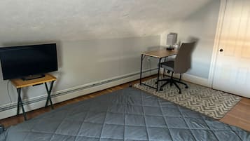 Quarto pequeno e acolhedor com televisão sobre suporte de madeira, escrivaninha com cadeira e tapete estampado no chão.