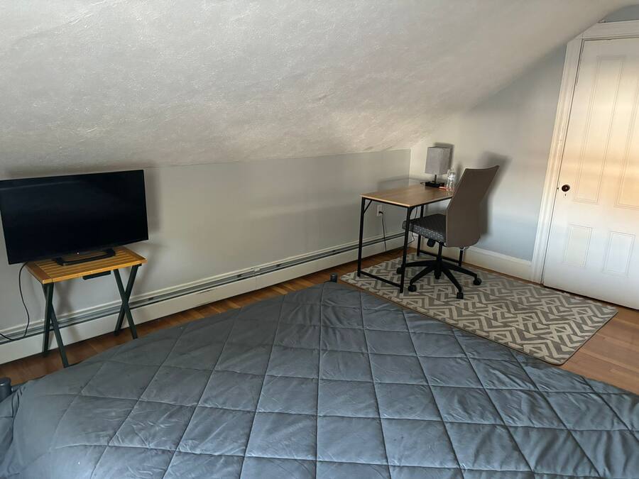 Quarto pequeno e acolhedor com televisão sobre suporte de madeira, escrivaninha com cadeira e tapete estampado no chão.