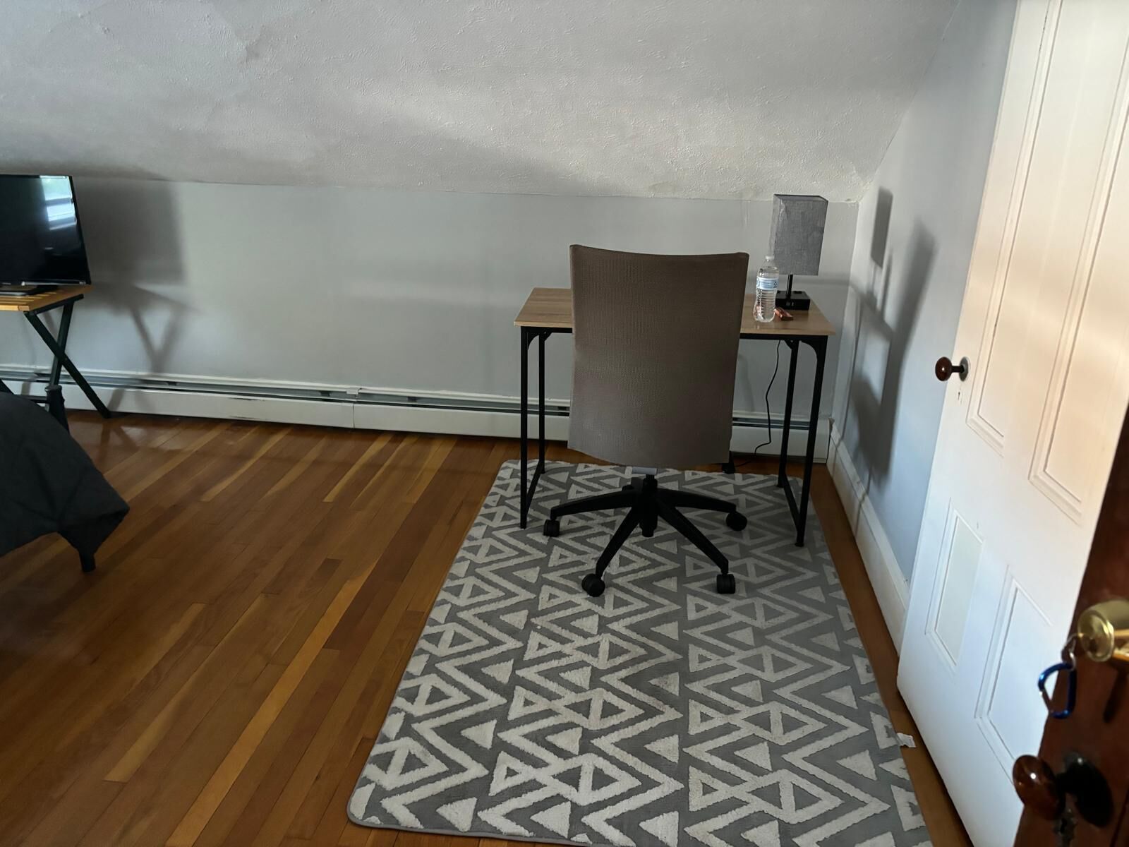 A room with a wooden floor, a patterned rug, a desk with a chair, and a closed door.