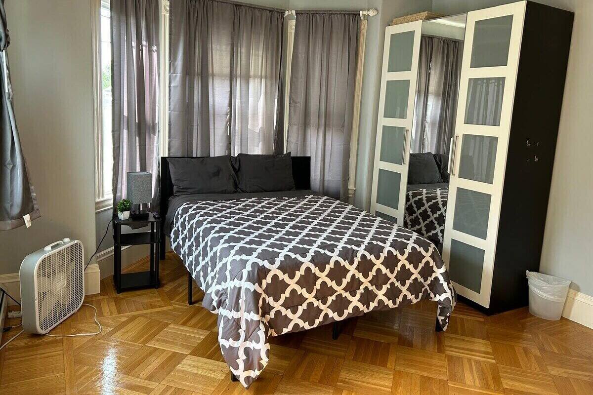 A bedroom with a bed, a wardrobe, a nightstand, a fan, and a window with curtains.