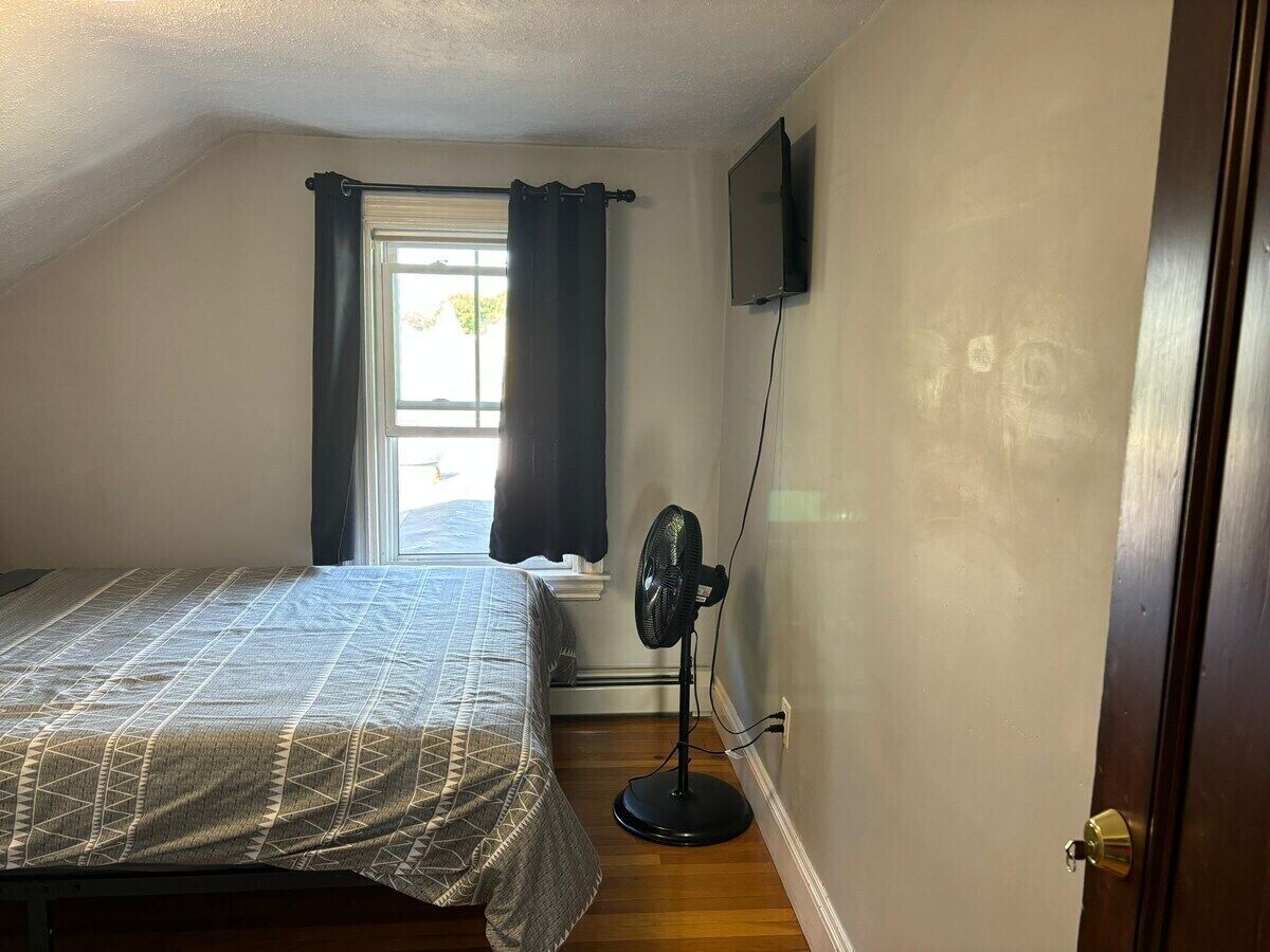 A bedroom with a bed, a standing fan, a window with curtains, and a wall-mounted TV.