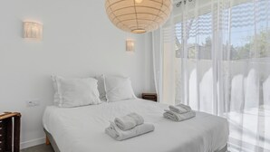 Quarto com uma cama, duas mesinhas de cabeceira, uma luminária na parede e cortinas leves.