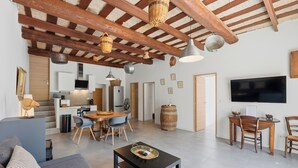 Uma sala ampla com teto de madeira, mesa de jantar com cadeiras, sofá, televisão e cozinha ao fundo.