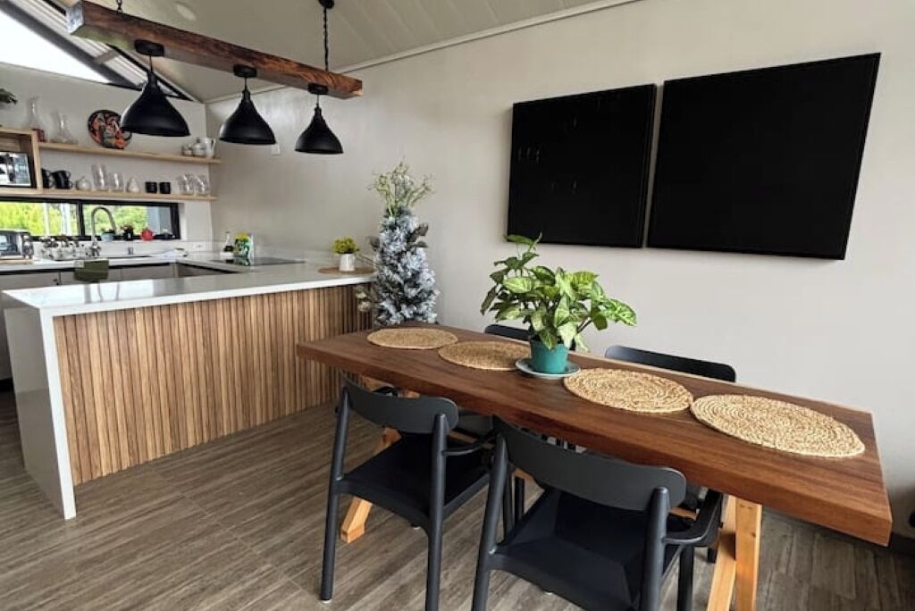 Uma cozinha moderna com uma ilha de madeira, luminárias pendentes pretas e uma área de jantar com mesa e cadeiras de madeira, ambas pretas.