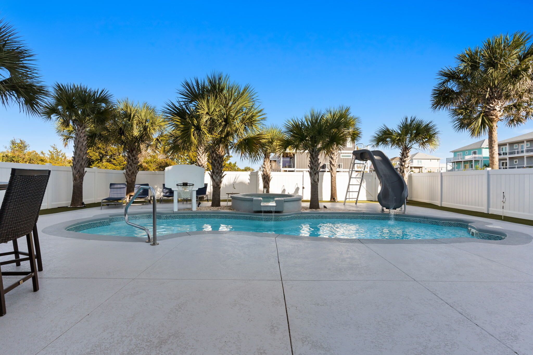 A swimming pool with lounge chairs and palm trees, surrounded by a white fence and buildings in the background.