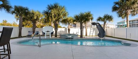 A swimming pool with lounge chairs and palm trees, surrounded by a white fence and buildings in the background.