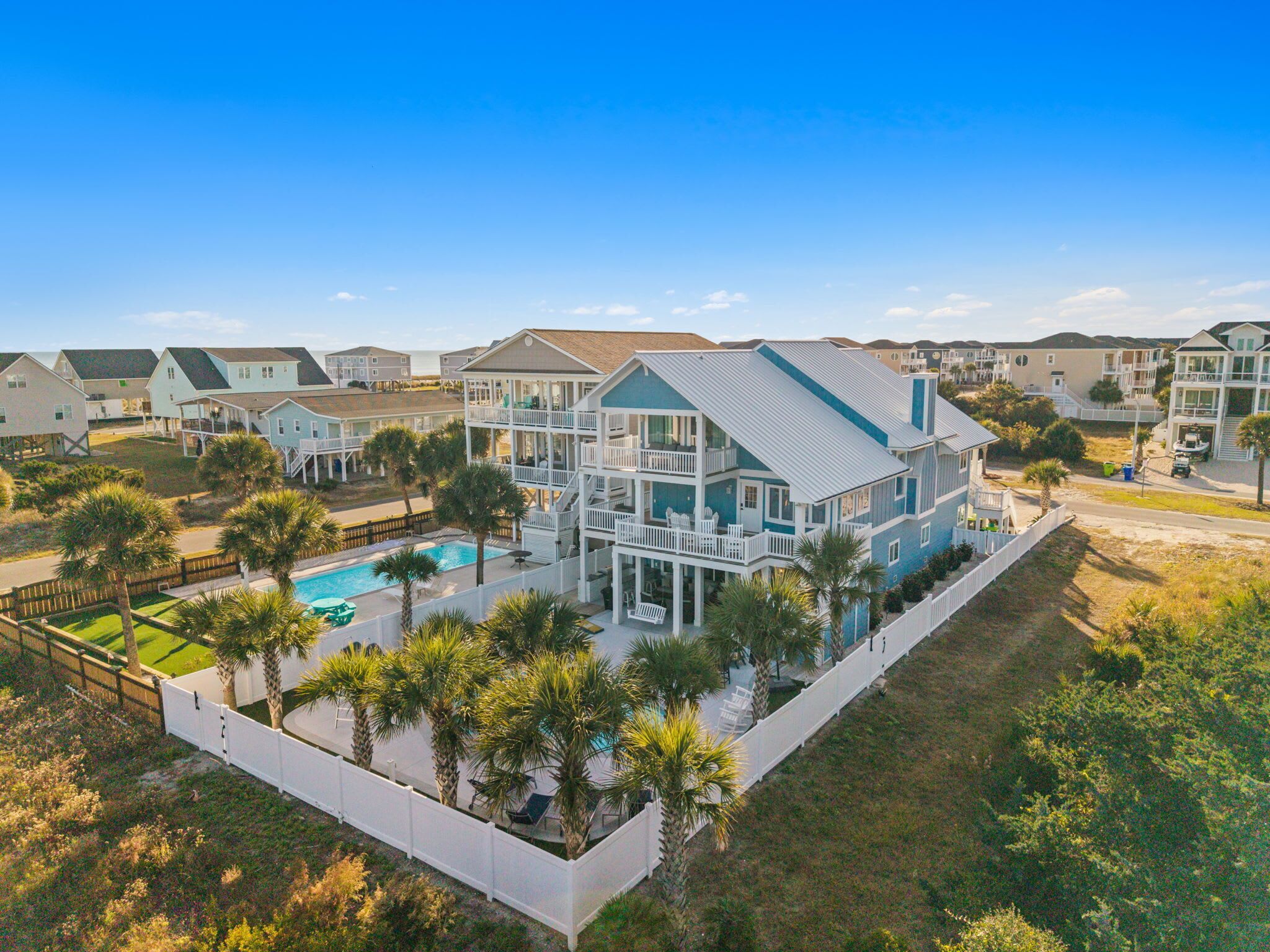 A large, multi-story house with a swimming pool, surrounded by palm trees and a white fence.