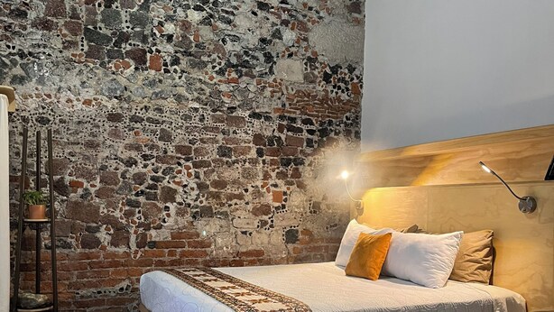 Ein Zimmer mit Backsteinwand, einem Bett mit Kissen, einer Kaffeetasse auf einem Tisch und einer Pendelleuchte.