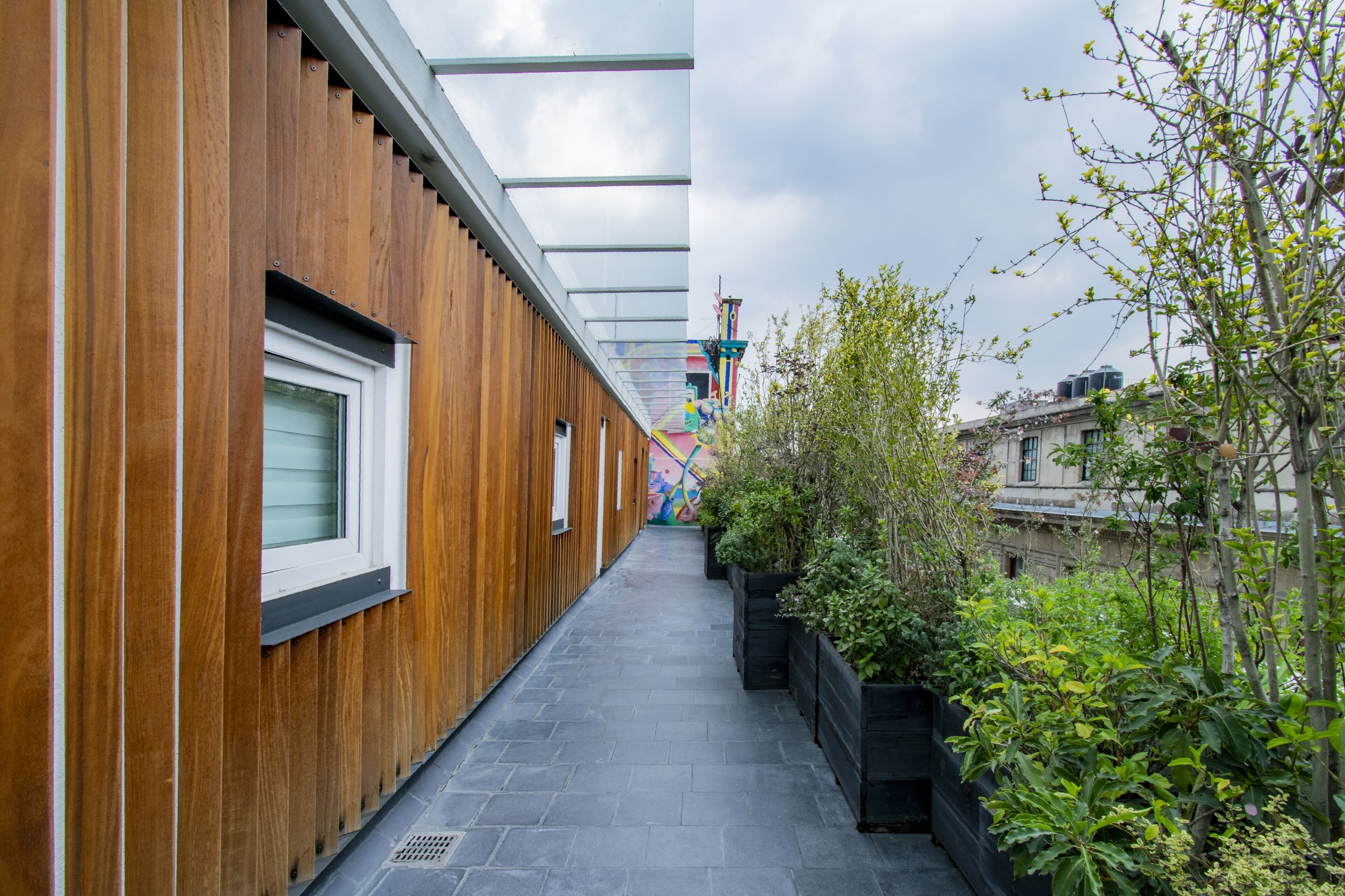 A narrow outdoor corridor with wooden paneling on one side and a black tiled floor. There are planters with greenery along the right side.