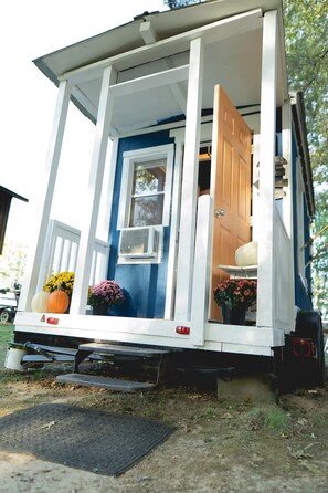 A mobile home with a porch, a sliding door, and a window with an air conditioning unit. - Charming Music City Tiny House (Ashland City)