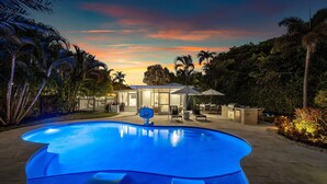 Una piscina con aro de baloncesto, rodeada de palmeras y un patio con mobiliario exterior.