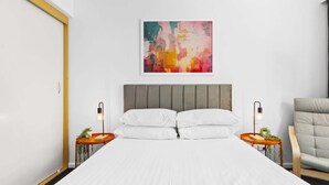 Quarto com cama, duas mesas de cabeceira com abajures, uma cadeira e um quadro abstrato colorido na parede.