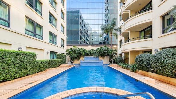 Piscina com bordas azulejadas, rodeada por sebes bem aparadas e um edifício com vários andares e varandas.
