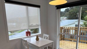 Een kamer met een witte tafel en stoelen, een raam met zonwering en uitzicht op een terras en buitenruimte.