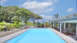 En swimmingpool med en overdækket patio, omgivet af frodig vegetation og et stort træ.