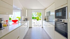 Une cuisine moderne avec des meubles blancs, un grand îlot central et une vue sur un jardin, accessible par une porte ouverte.